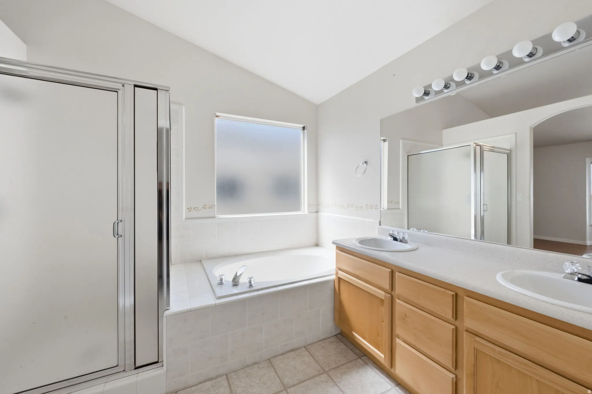 Bathroom featuring a dual vanity with light wood cabinetry and integrated sinks, a built-in tub with tile surround, a frosted glass window, and a separate shower with a glass enclosure