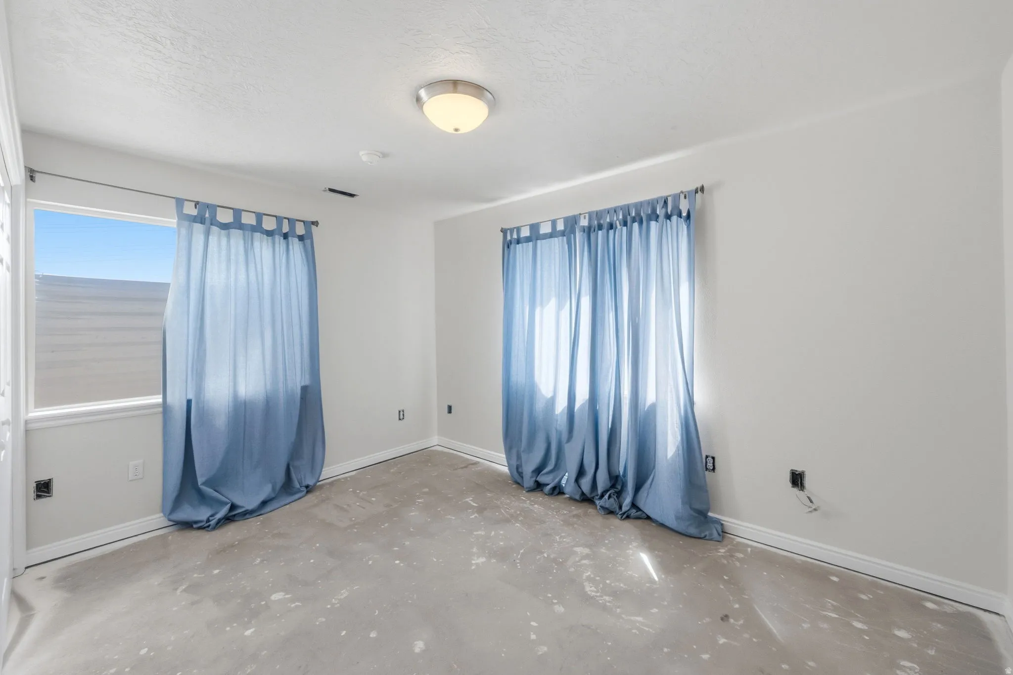 Room featuring two windows with blue curtains, flush mount lighting, textured ceiling, and white baseboards