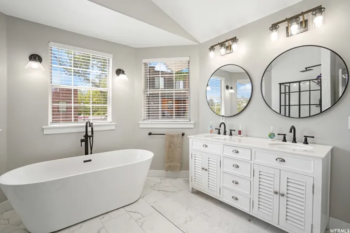 Contemporary bathroom featuring a freestanding soaking tub with a floor-mounted matte black faucet, a dual vanity with white cabinetry and louvered doors, a white countertop, and two large circular mirrors
