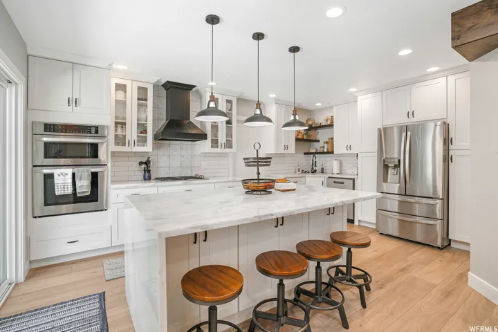 Bright kitchen featuring a waterfall island with stone countertop, stainless steel appliances including a double oven and refrigerator, white cabinetry, and wood-finish flooring
