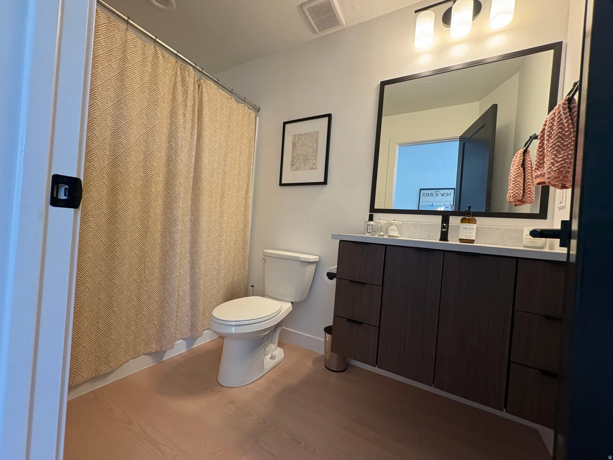 Bathroom featuring a floating wood-finish vanity with a light countertop, a rectangular framed mirror, a three-light fixture, and light wood-finish flooring