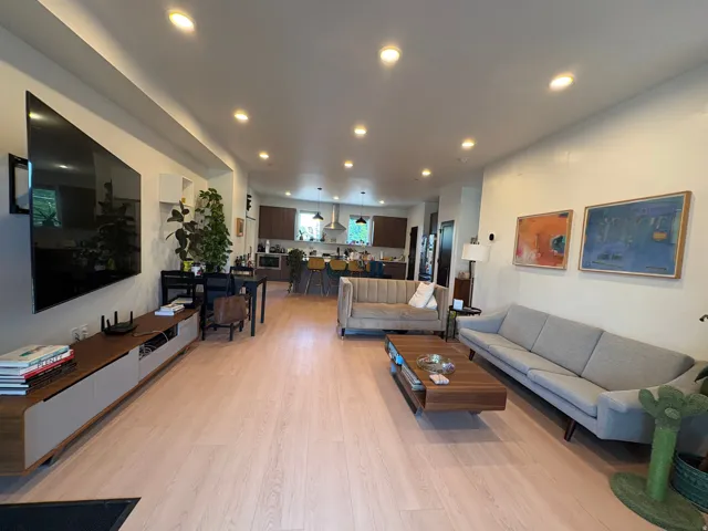 Open-concept living space featuring light wood-finish flooring, recessed lighting, and a modern kitchen visible in the background