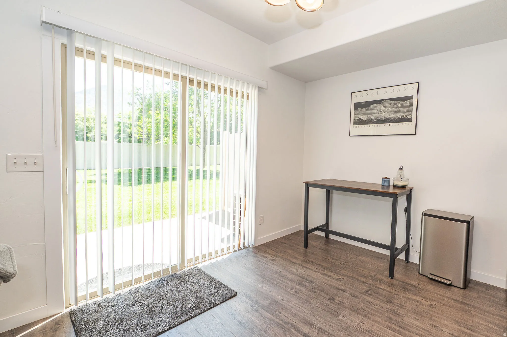 Sliding glass door with vertical blinds opening to a concrete patio and a fenced yard with mature trees