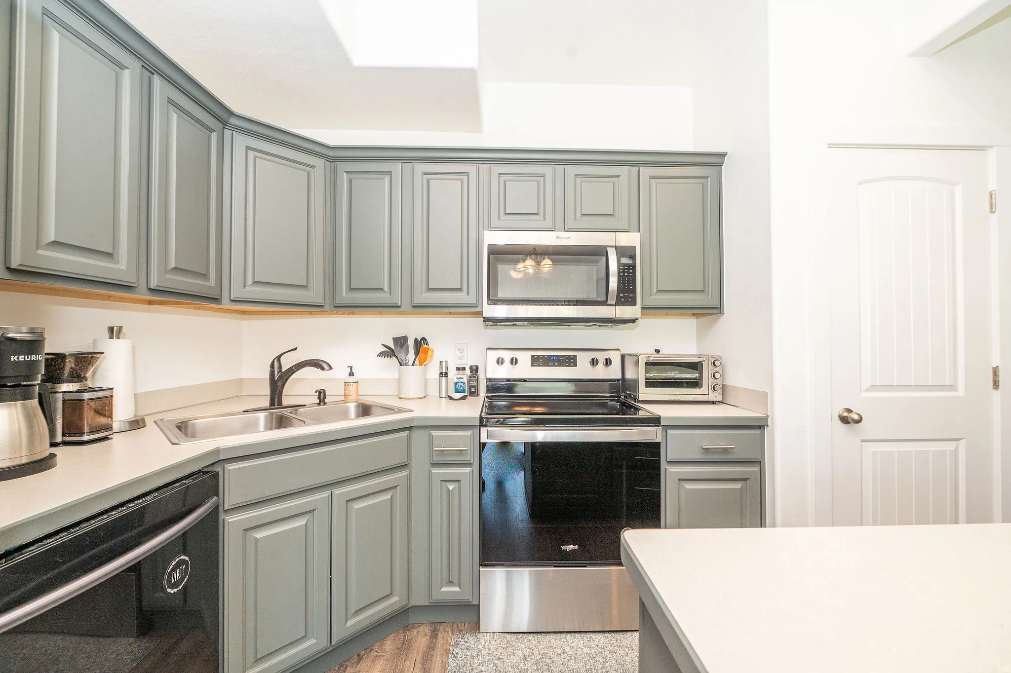 Kitchen featuring gray shaker-style cabinetry, light-toned countertops, a stainless steel double-basin sink with a matte black faucet, and stainless steel appliances including an oven range and microwave
