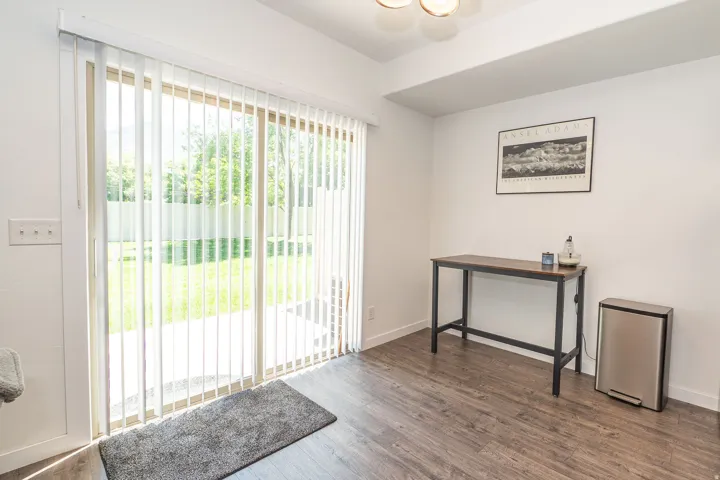 Sliding glass door with vertical blinds opening to a concrete patio and a fenced yard with mature trees