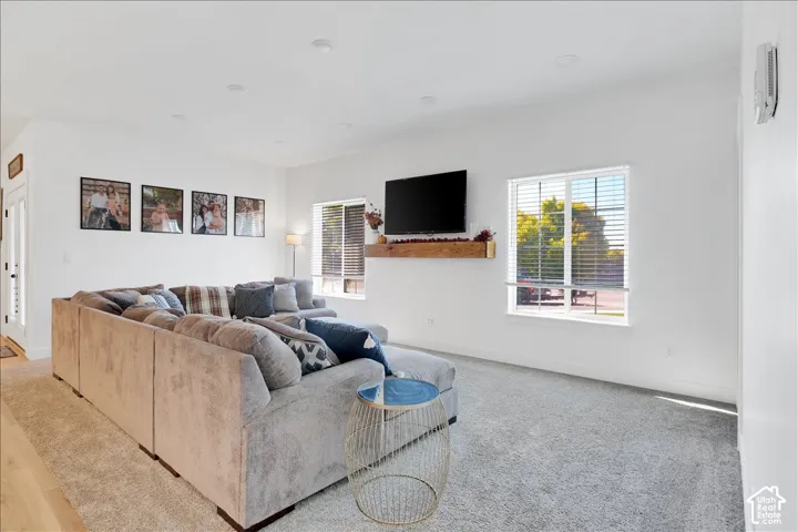 Spacious living area featuring light gray carpeting, recessed ceiling lighting, two windows with blinds, a wall-mounted television, and a wood-finish floating shelf