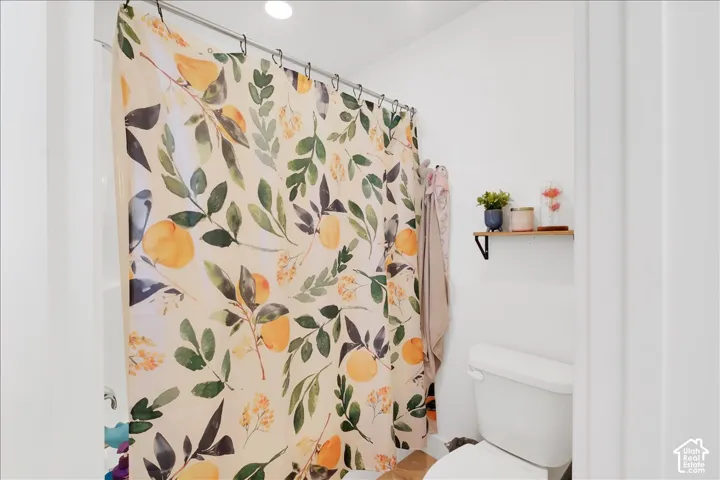 Bathroom featuring a white porcelain toilet, a white bathtub with an integrated shower, a chrome shower curtain rod, a recessed ceiling light, and a wall-mounted wood-finish shelf with black metal brackets