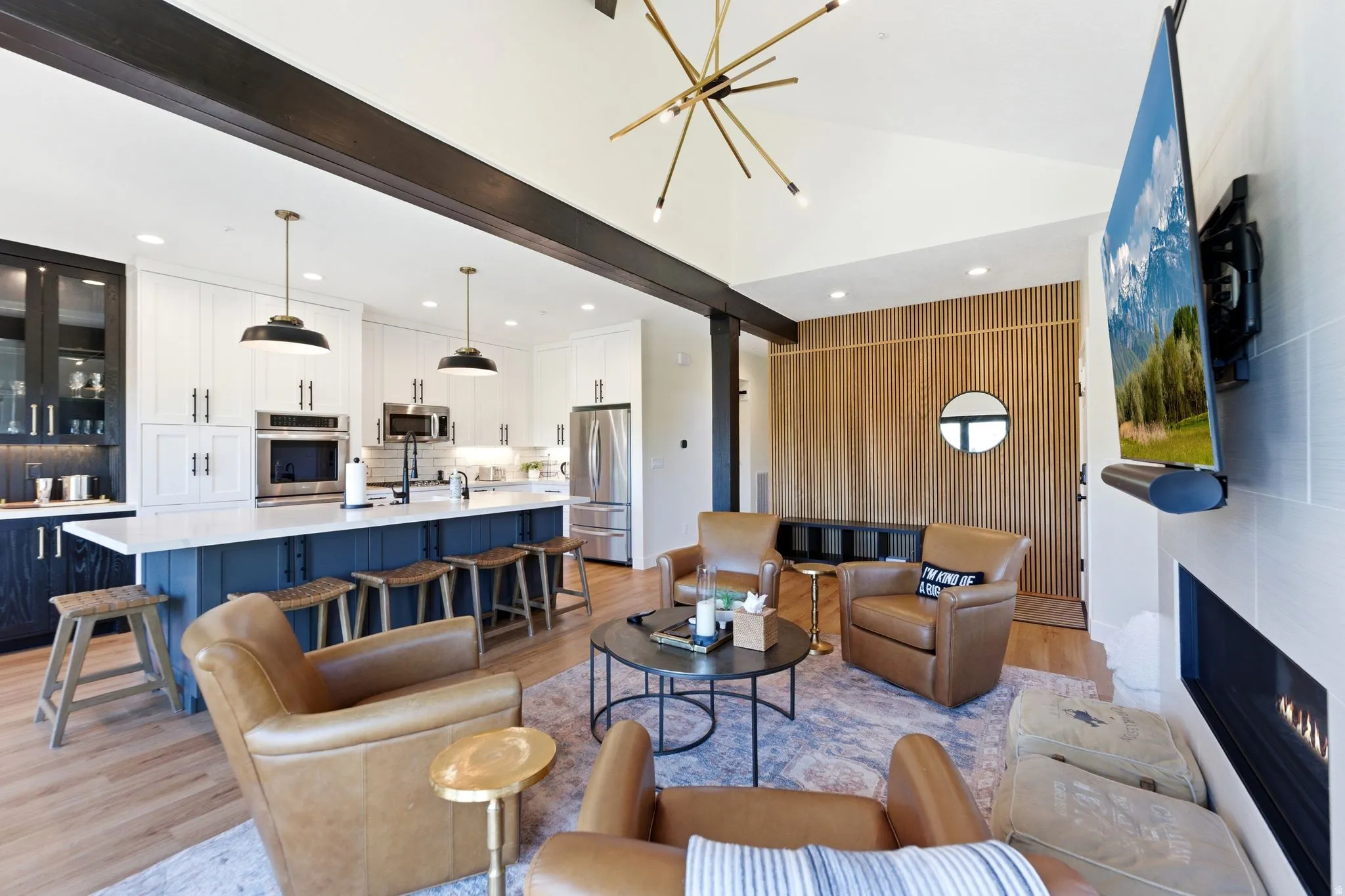 Open-concept living space featuring a vaulted ceiling with exposed beams, wood-finish flooring, a linear fireplace, custom kitchen cabinetry with a central island, and a distinctive wood slat accent wall