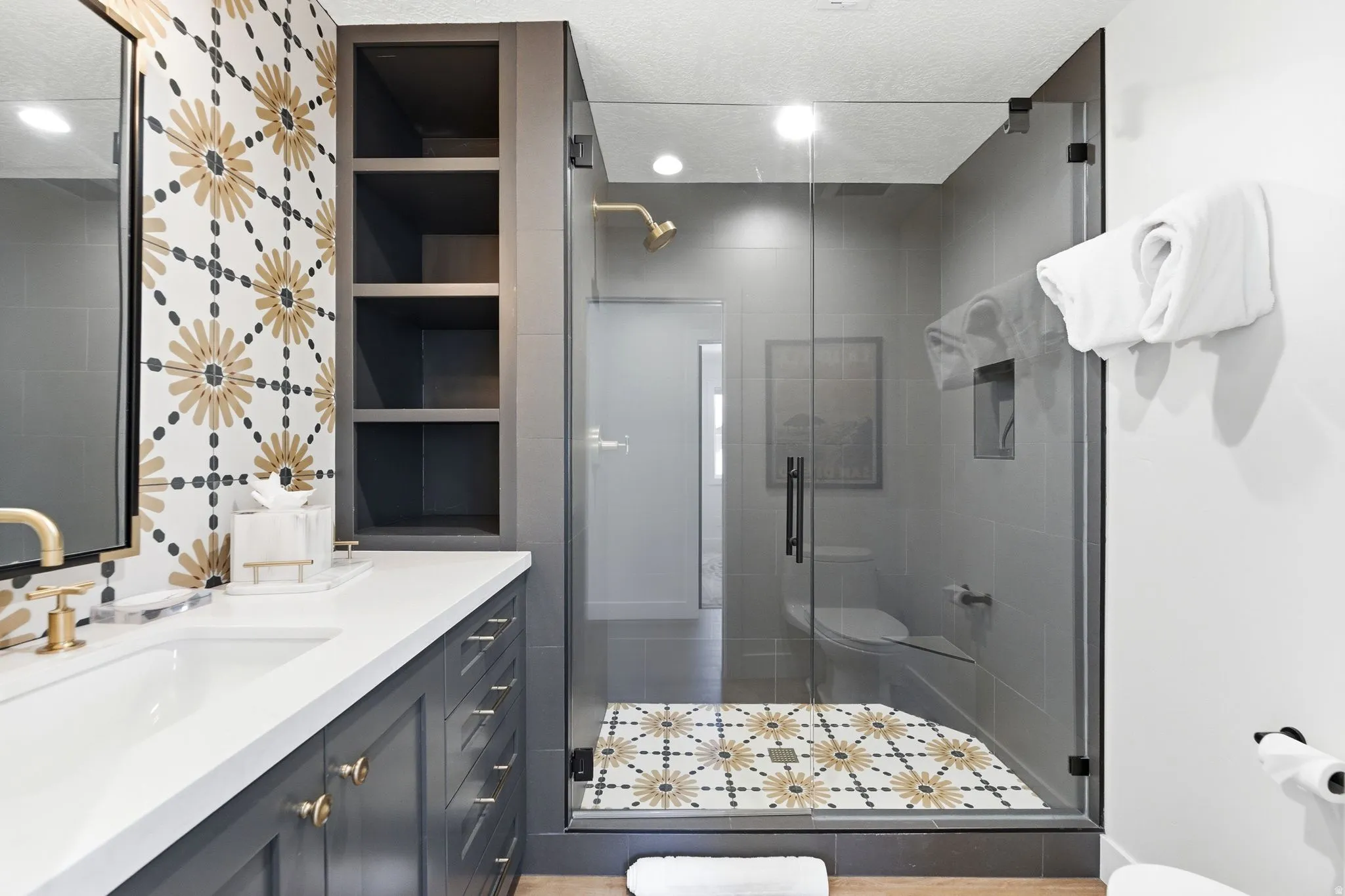 Bathroom featuring a glass-enclosed shower with decorative floor tiling, a vanity with dual sinks, a quartz countertop, and gold-finish fixtures