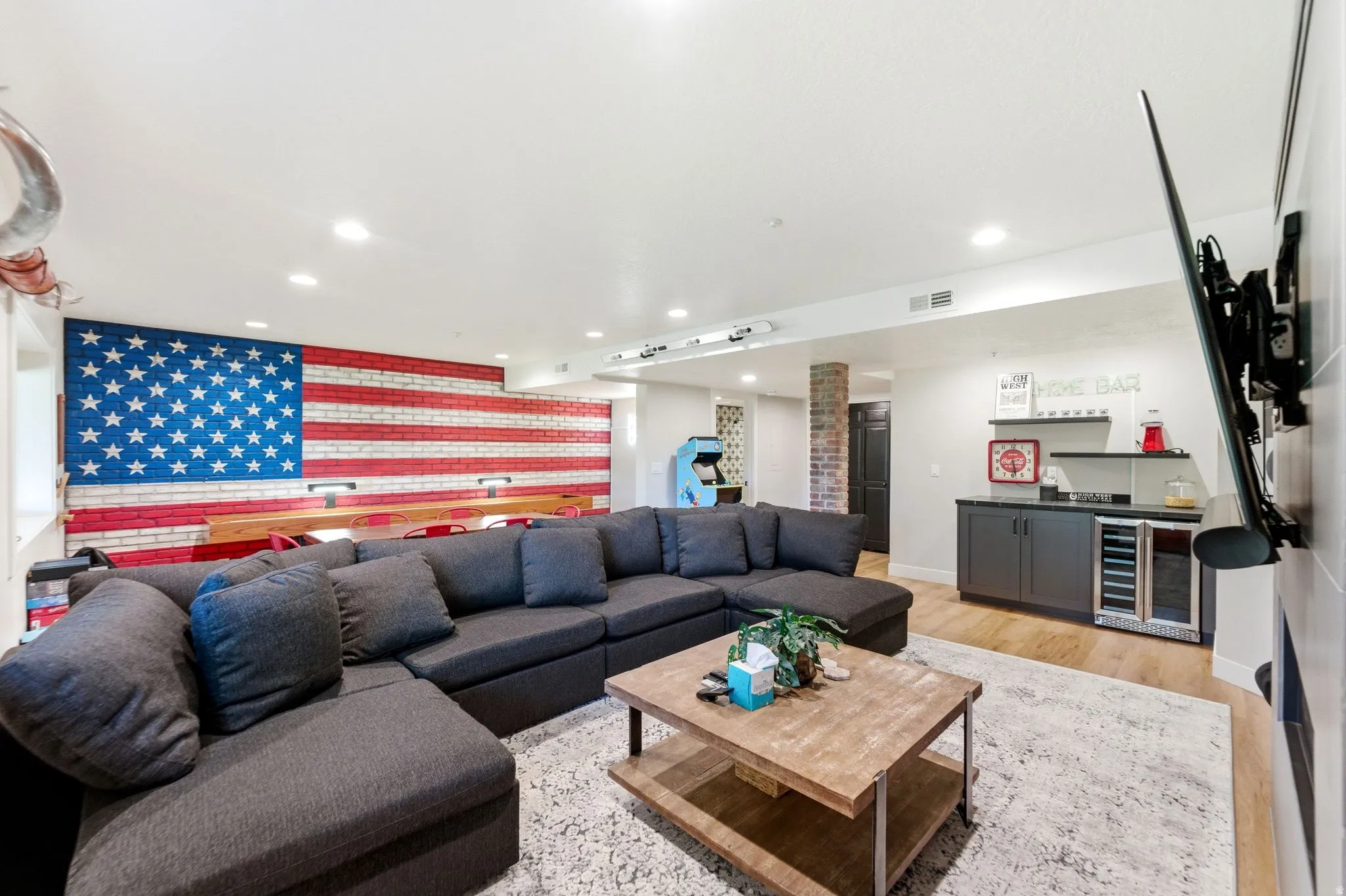 Spacious entertainment room featuring a full wall brick accent, recessed lighting, wood-finish flooring, and a built-in wet bar with a beverage cooler and dark cabinetry