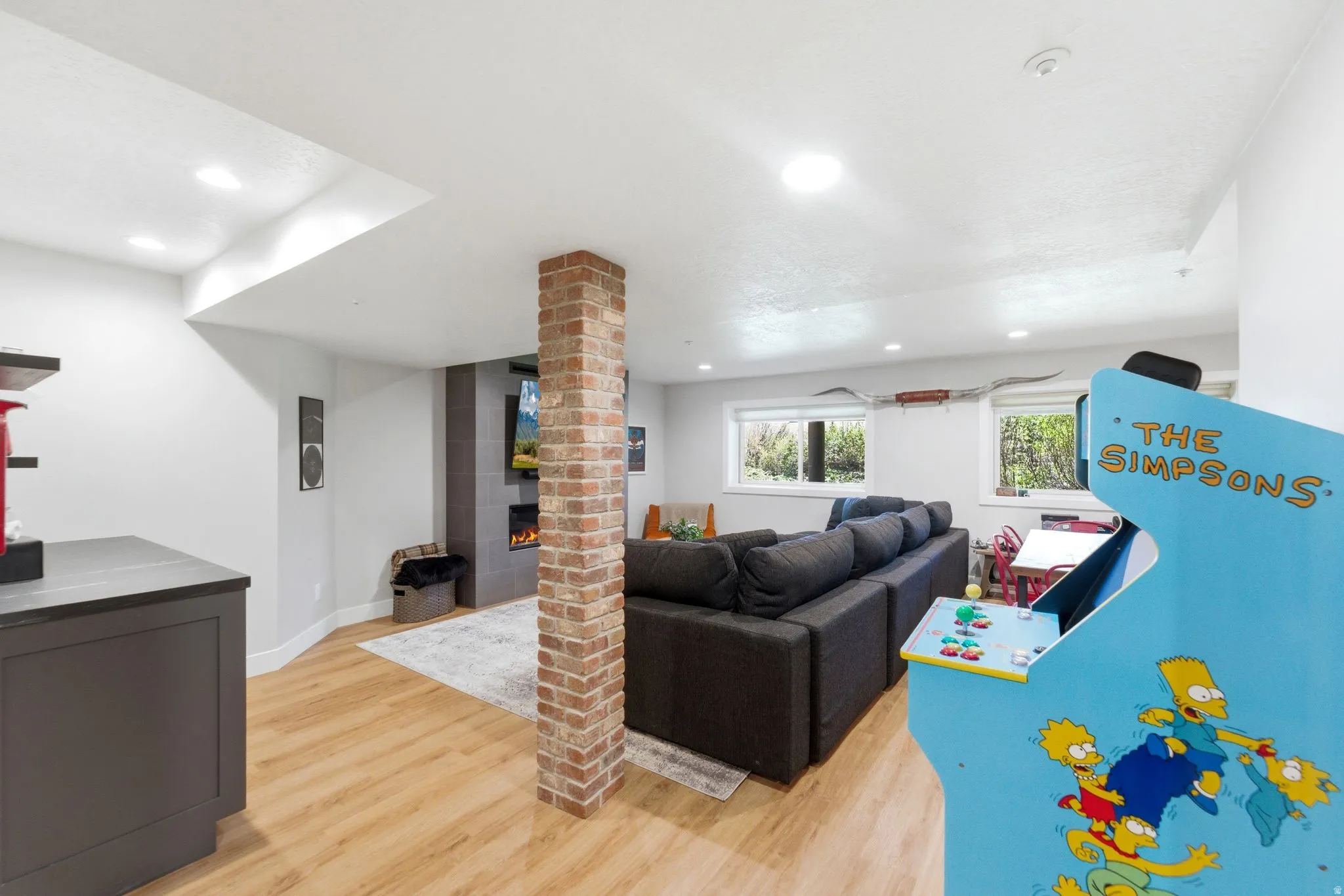 Finished lower-level space featuring wood-finish flooring, a brick column, a fireplace with a dark tile surround, and recessed lighting