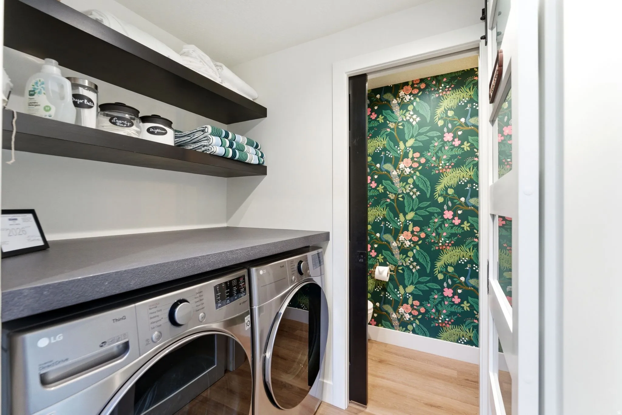 Laundry area featuring a dark gray countertop, stacked front-load machines, and two dark wood-finish floating shelves