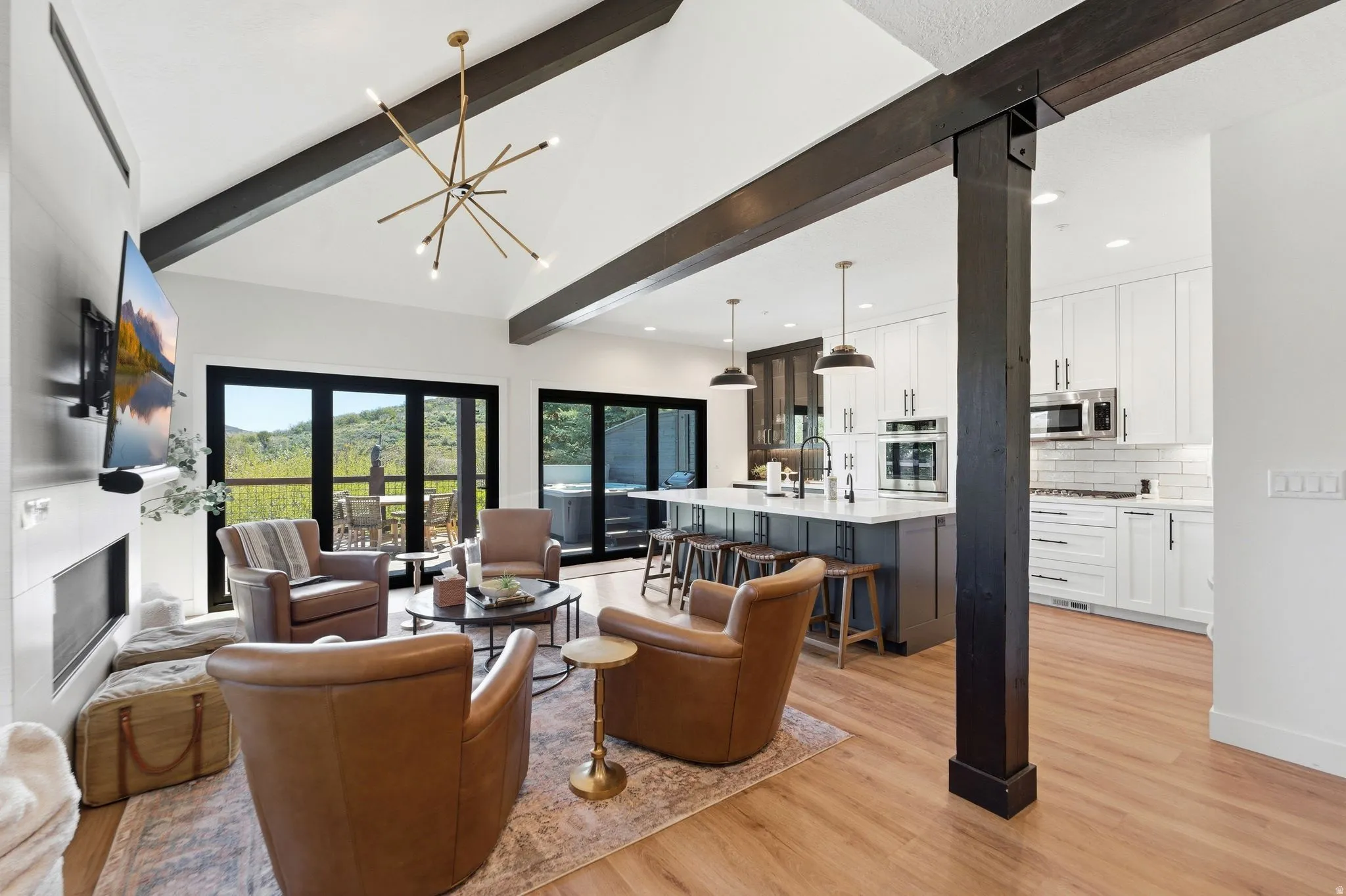 Open-concept living space featuring vaulted ceilings with exposed beams, wood-finish flooring, a contemporary fireplace, a modern kitchen with a large island, and expansive glass doors opening to the exterior
