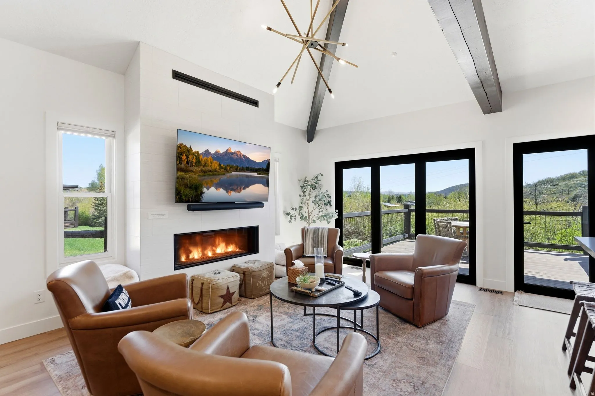 Living area featuring a modern linear fireplace, high vaulted ceilings with exposed beams, wood-finish flooring, and multiple glass doors opening to an outdoor deck