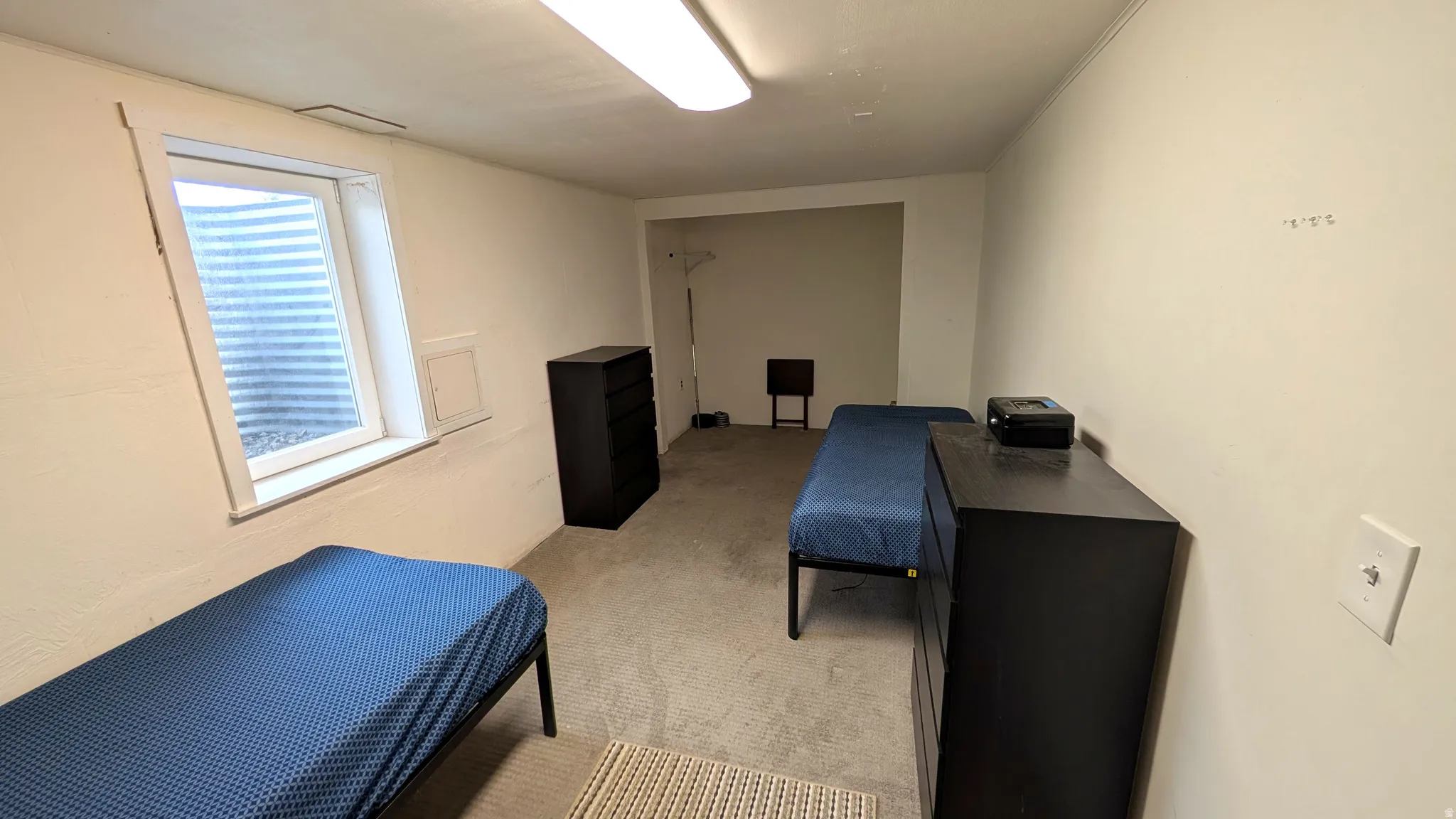 Carpeted room featuring a white-framed egress window, light-colored walls, and overhead fluorescent lighting