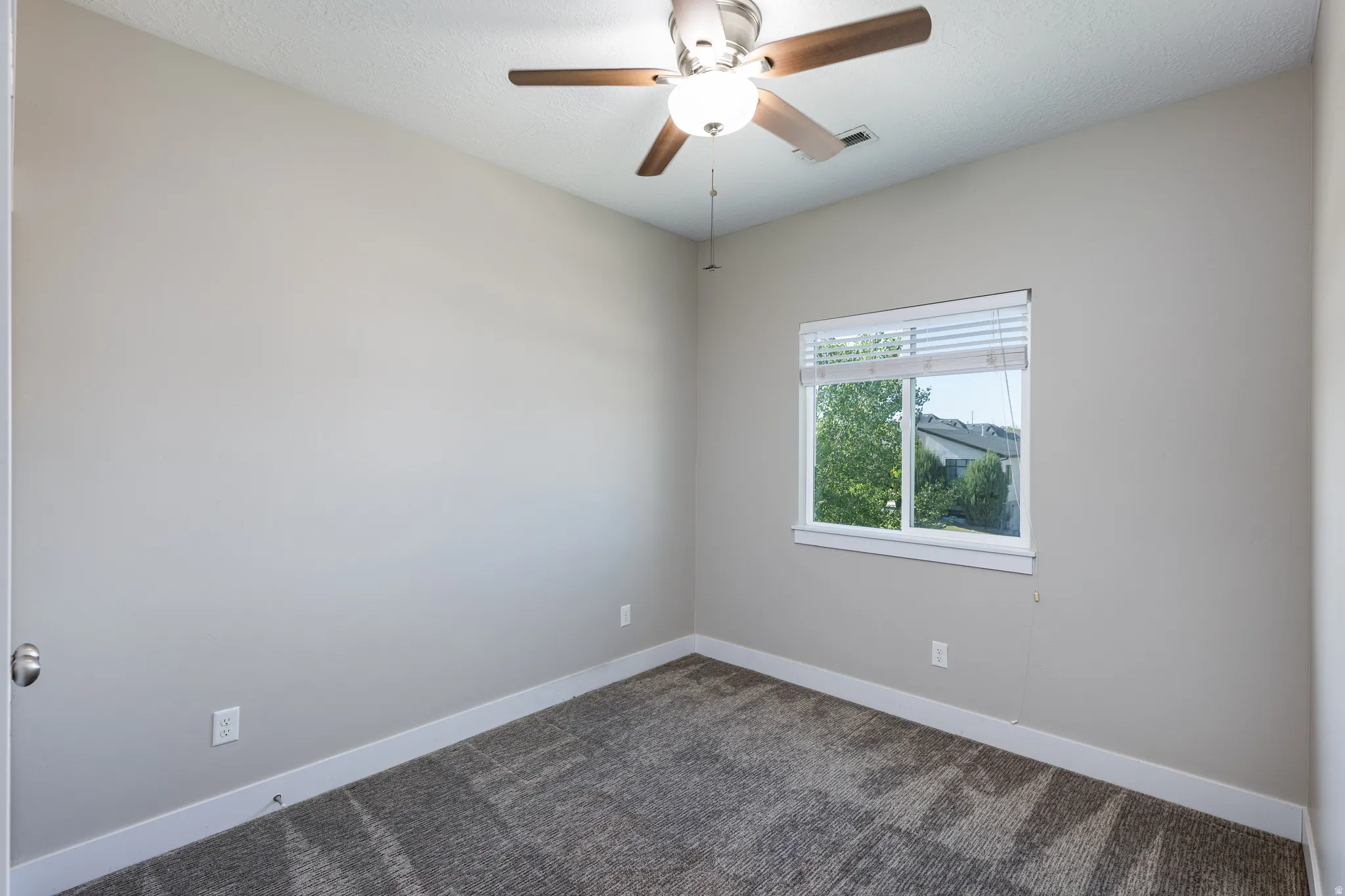 Neutral-toned interior room featuring patterned carpet flooring, a window with blinds, a ceiling fan with light fixture, and white baseboards