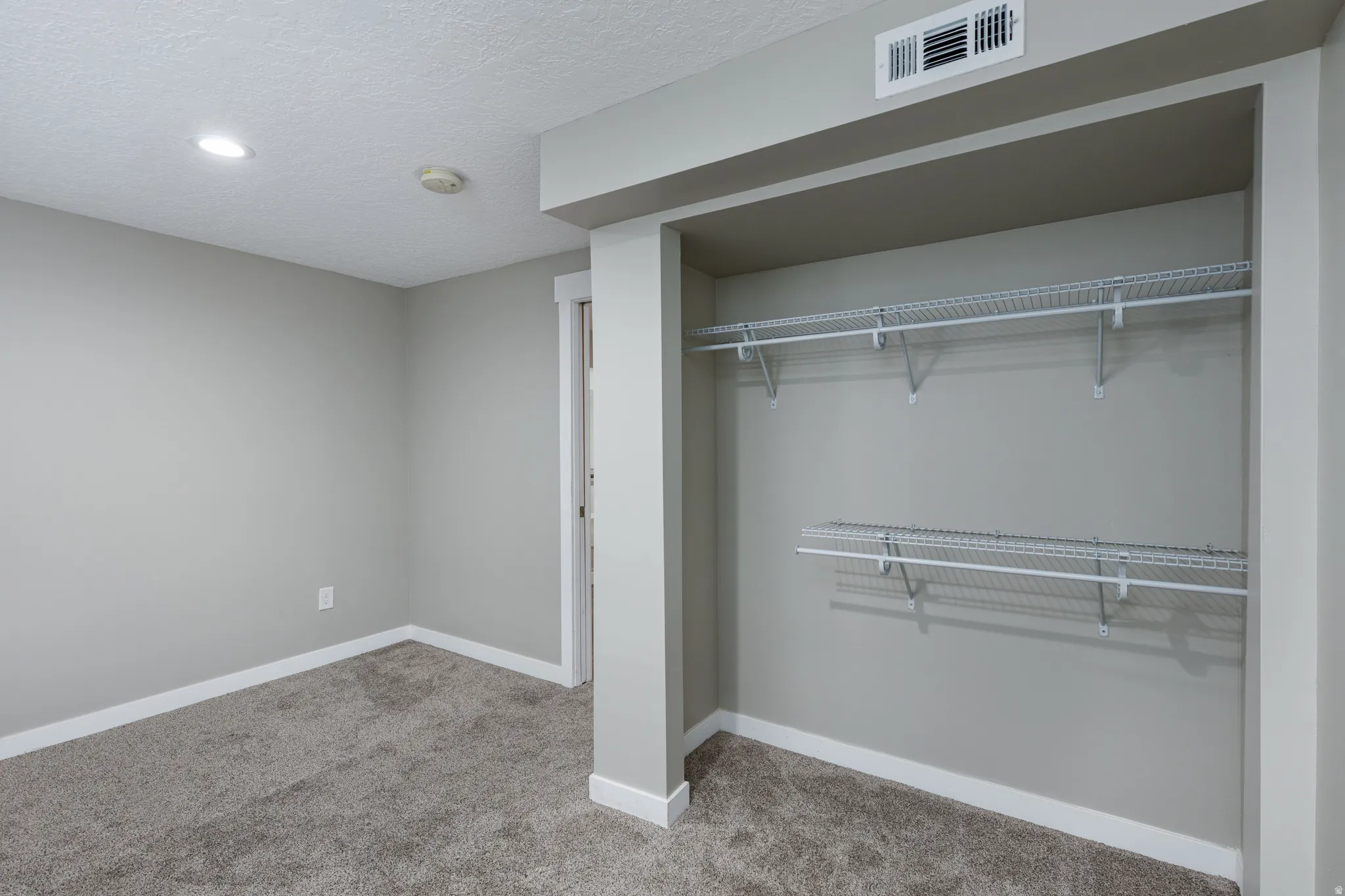 Recessed lighting illuminates a space featuring wire shelving, neutral carpeting, light gray walls, and white baseboards