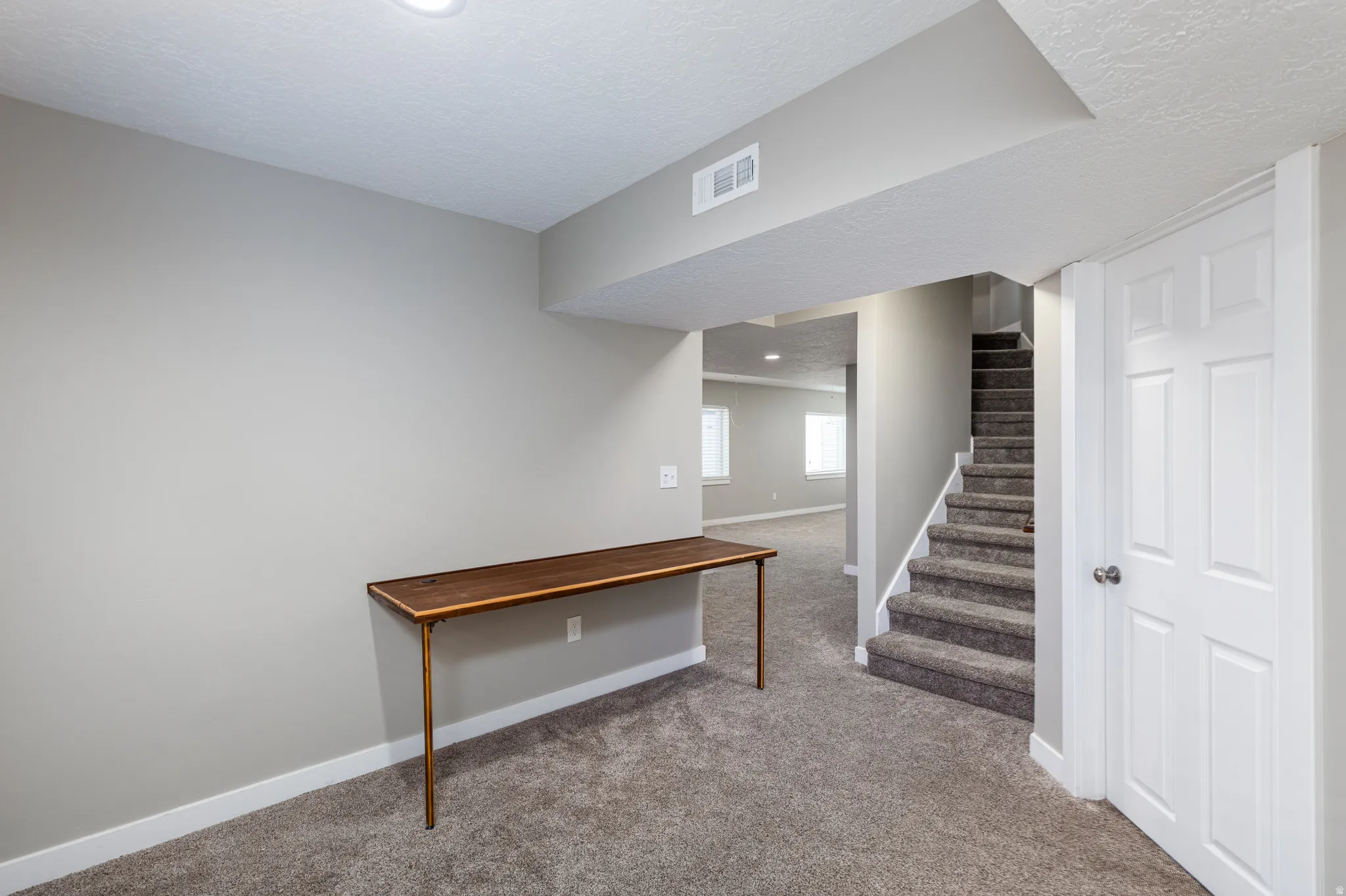 Lower level space featuring neutral gray wall paint, gray carpet flooring, a built-in wood-finish console table, and a staircase with white risers and gray carpeted treads