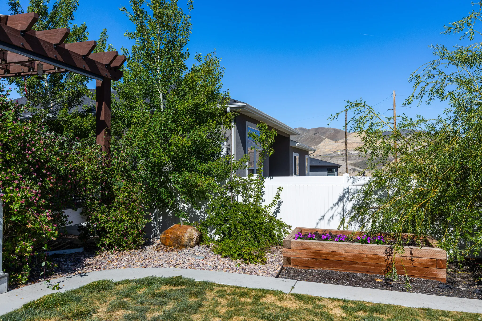 Wood pergola structure overlooking a landscaped yard featuring a wooden planter box with purple flowers, a rock garden, and a white privacy fence