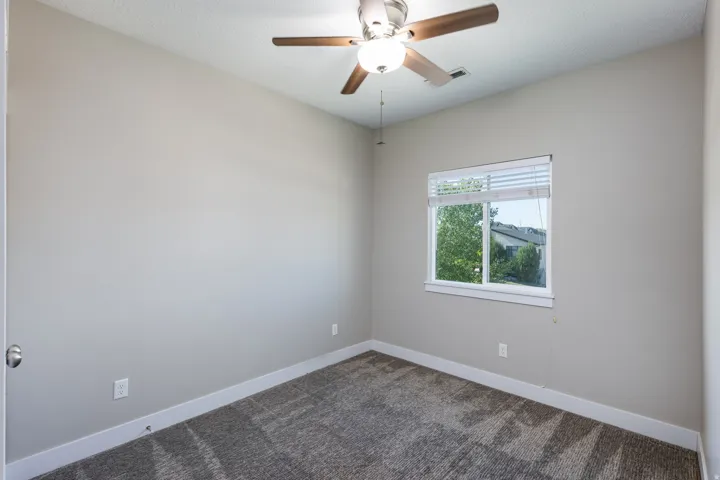 Neutral-toned interior room featuring patterned carpet flooring, a window with blinds, a ceiling fan with light fixture, and white baseboards