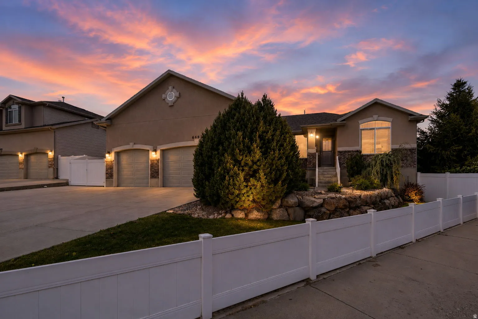 Stucco and stone facade home featuring a multi-car driveway, arched garage doors, established landscaping, and a white perimeter fence