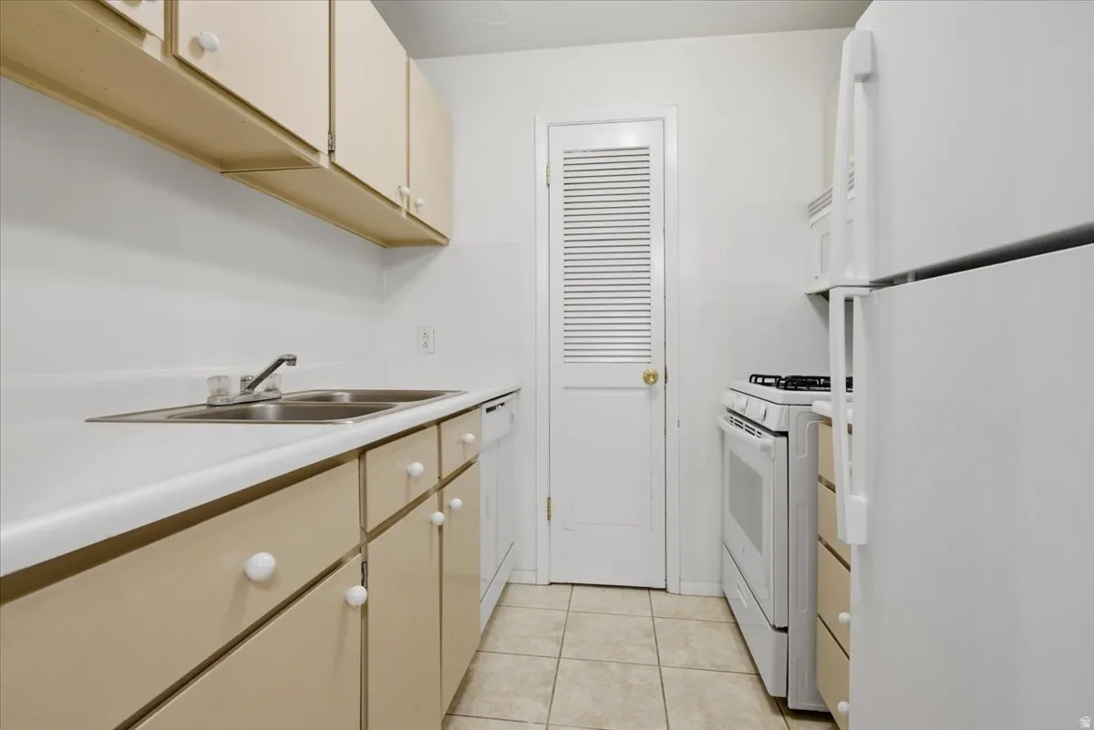 Kitchen featuring tan cabinetry, white countertops, a double basin stainless steel sink, tile flooring, and white appliances including a refrigerator, range, and built-in microwave