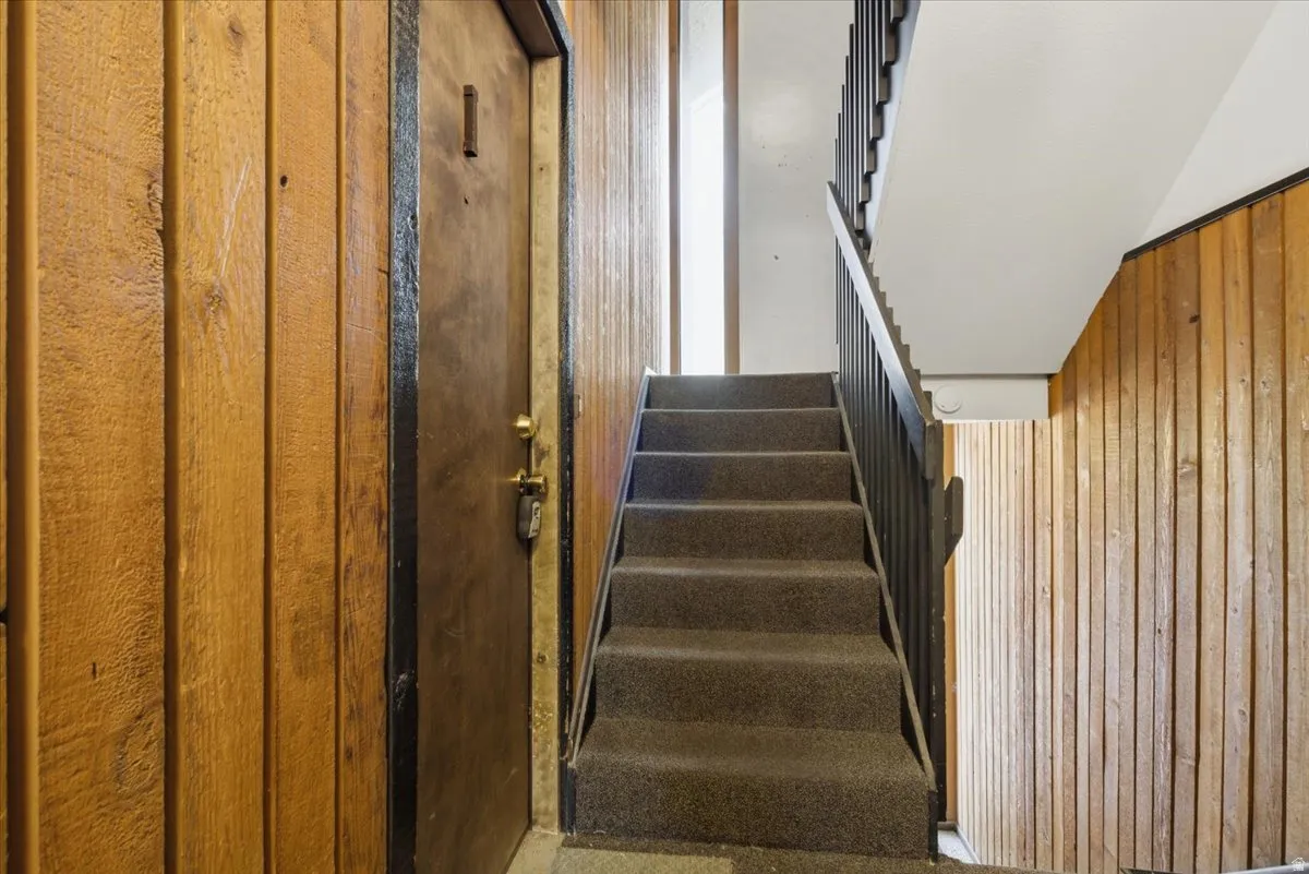 Carpeted staircase with dark metal handrail, vertical wood wall paneling, and a dark entry door with brass hardware