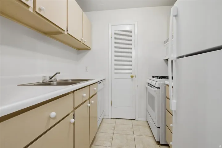 Kitchen featuring tan cabinetry, white countertops, a double basin stainless steel sink, tile flooring, and white appliances including a refrigerator, range, and built-in microwave