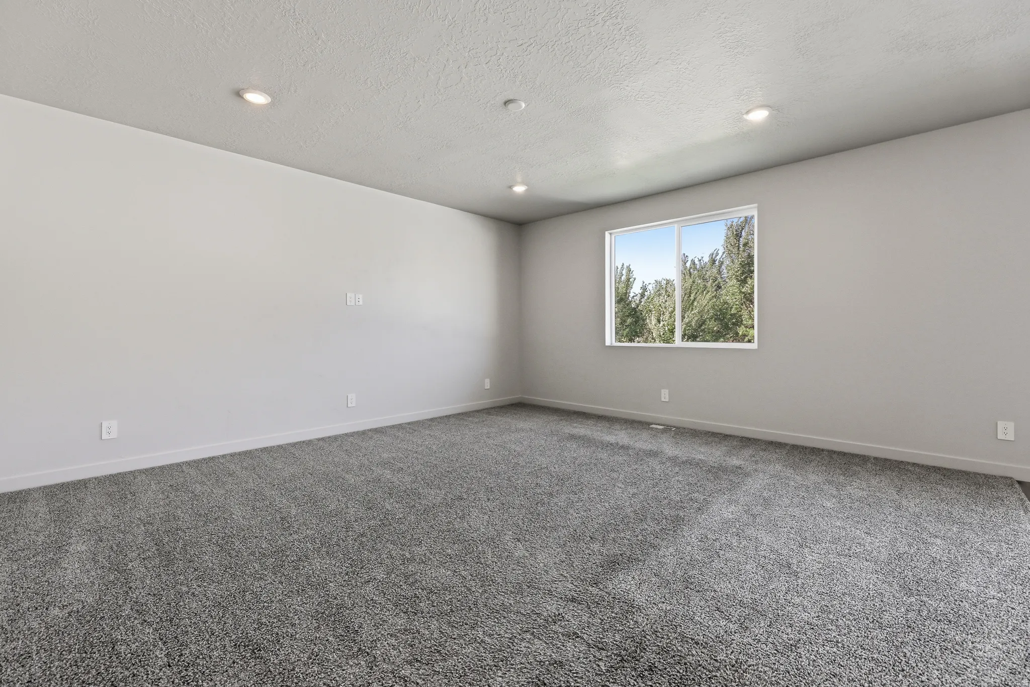 Recessed lighting, light gray walls, textured ceiling, gray carpeting, and a window with a white frame