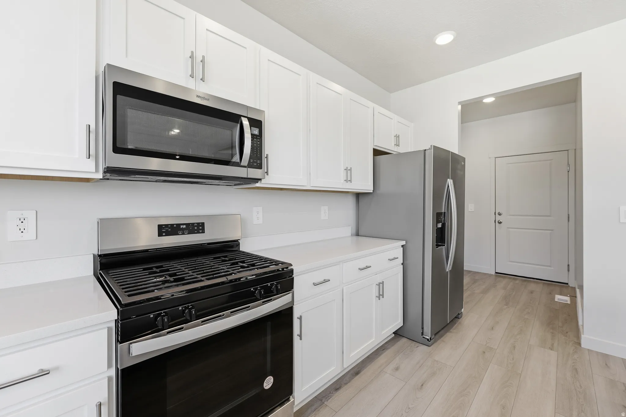 White shaker-style cabinetry, stainless steel appliances, light-toned wood-finish flooring, recessed ceiling lighting, and solid surface countertops