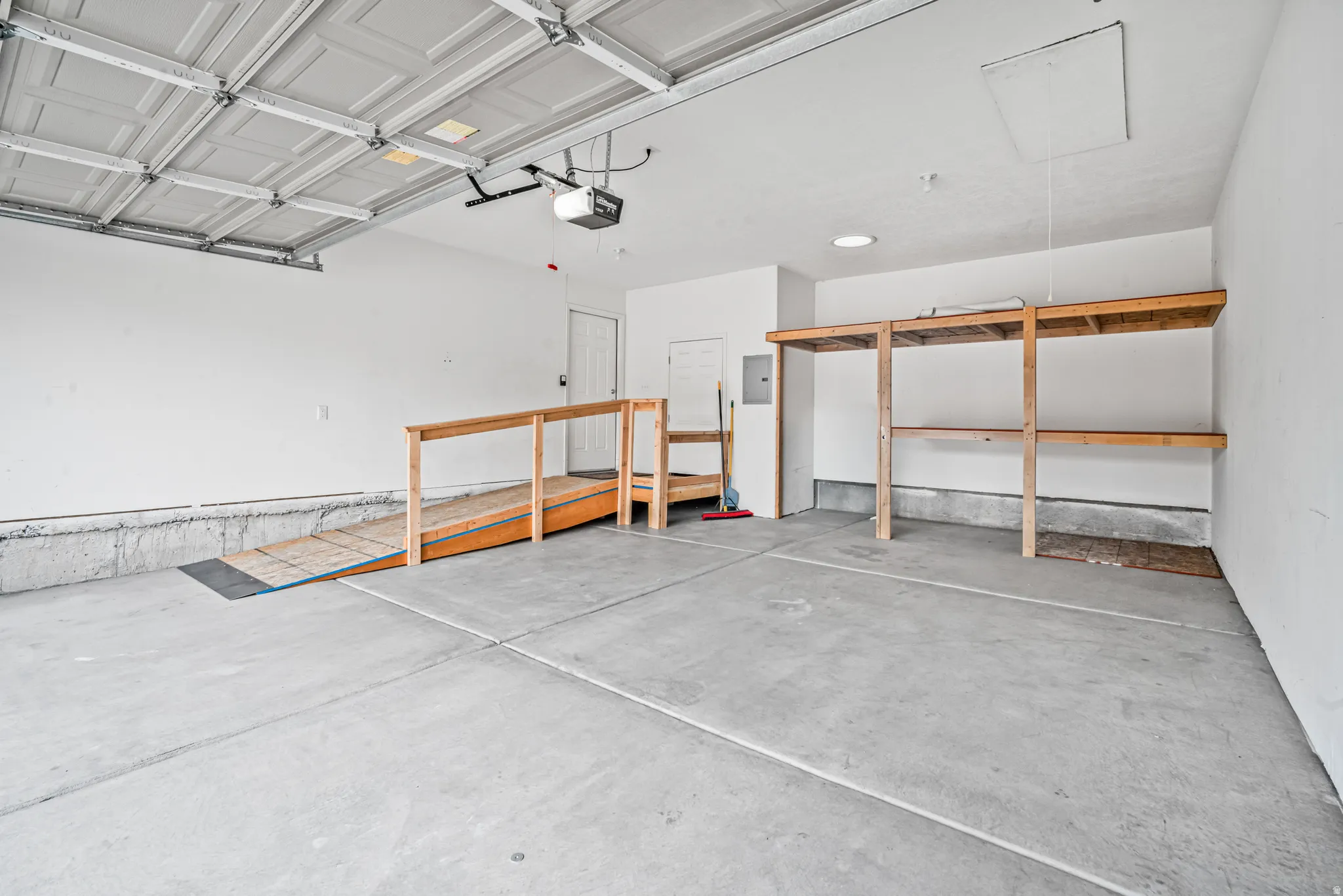 Spacious garage featuring a concrete floor, overhead garage door, built-in wooden ramp, and integrated wooden shelving.  Ramp can be easily removed