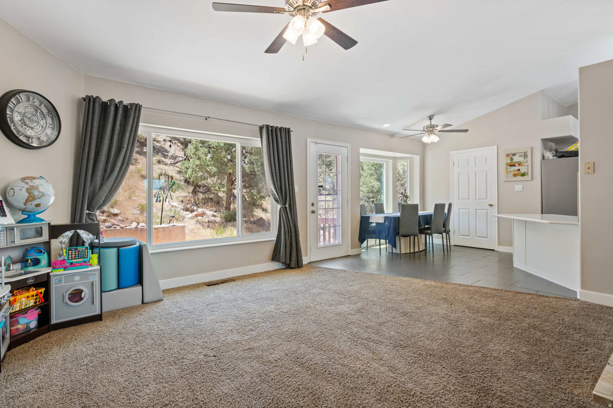 Spacious living area featuring ample natural light, vaulted ceilings, two ceiling fans, mixed flooring with carpet and tile, and a door leading to exterior space