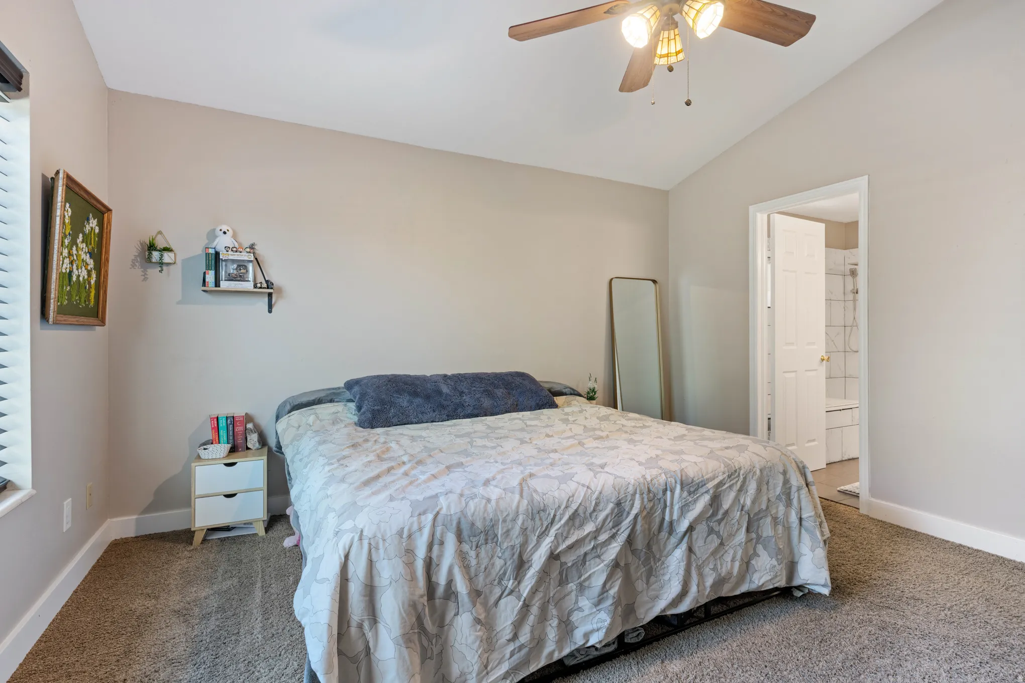 Carpeted bedroom featuring a vaulted ceiling, a ceiling fan with light fixture, window with blinds, and an entryway to an attached bathroom with a tiled shower