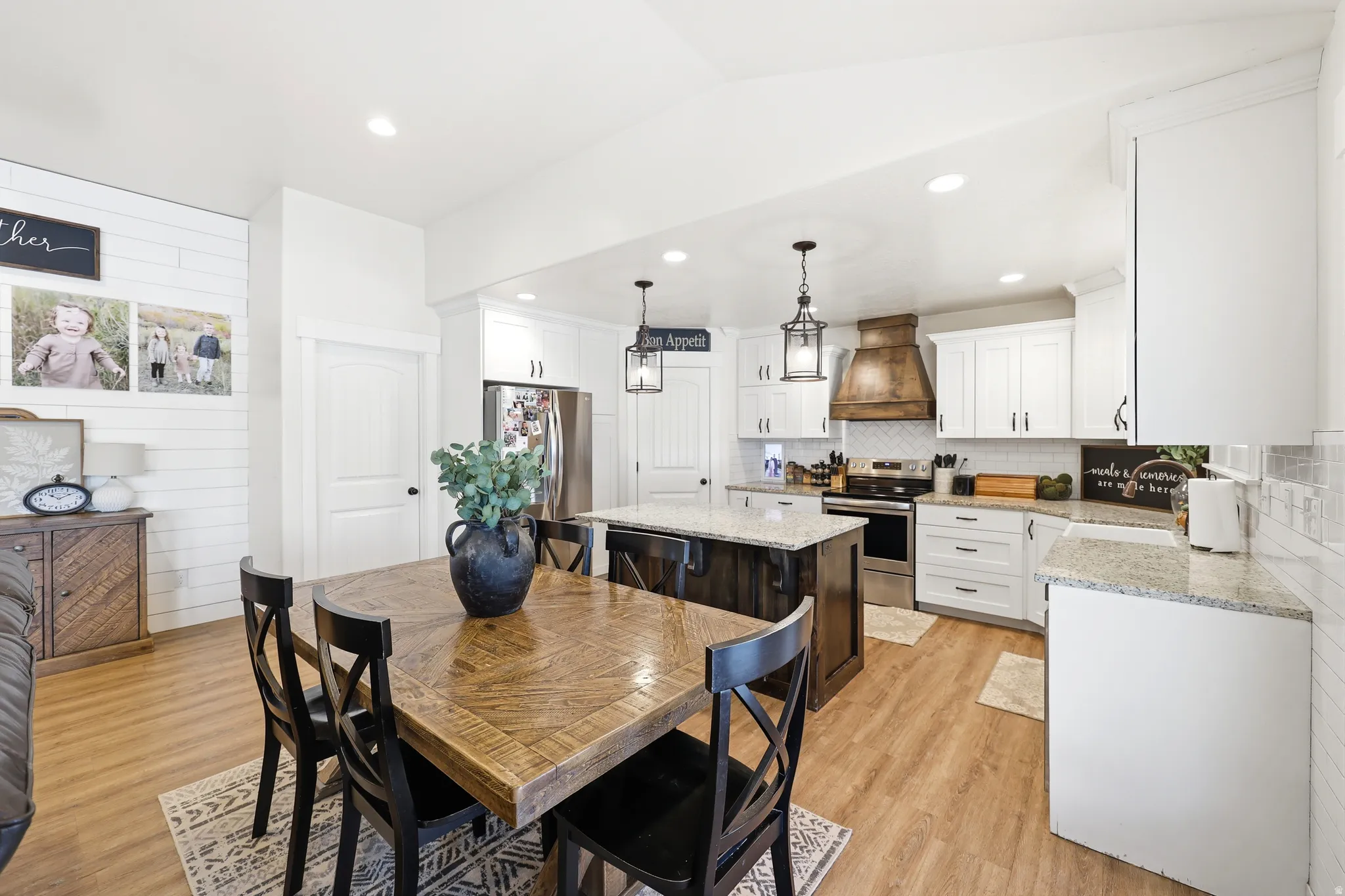 Open-concept kitchen and dining area featuring wood-finish flooring, white cabinetry, granite countertops, stainless steel appliances, and a rustic wood range hood