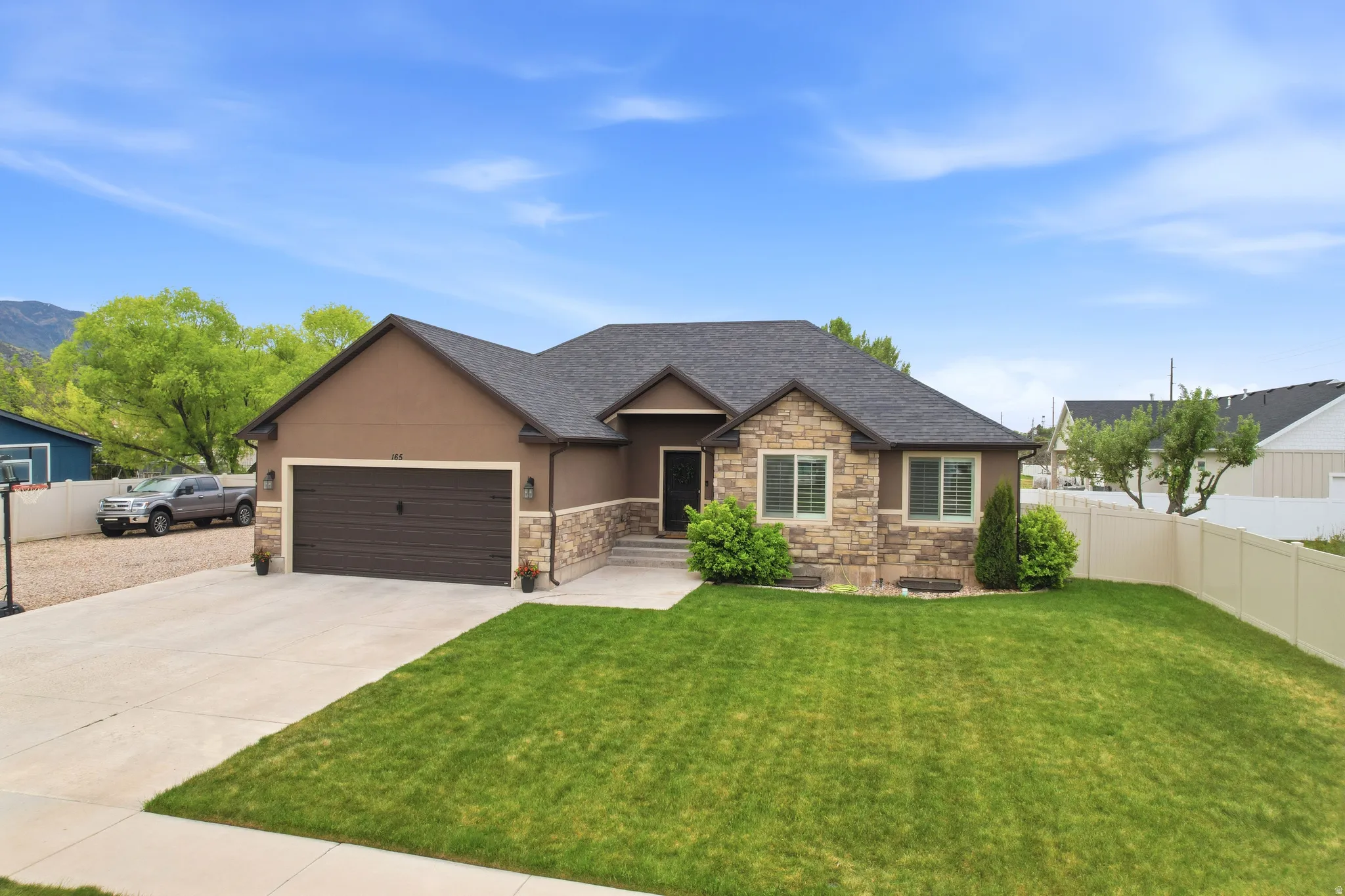 Front exterior featuring a stone and stucco facade, a dark shingle roof, an attached two-car garage with a dark wood-finish door, a concrete driveway, and a fenced backyard