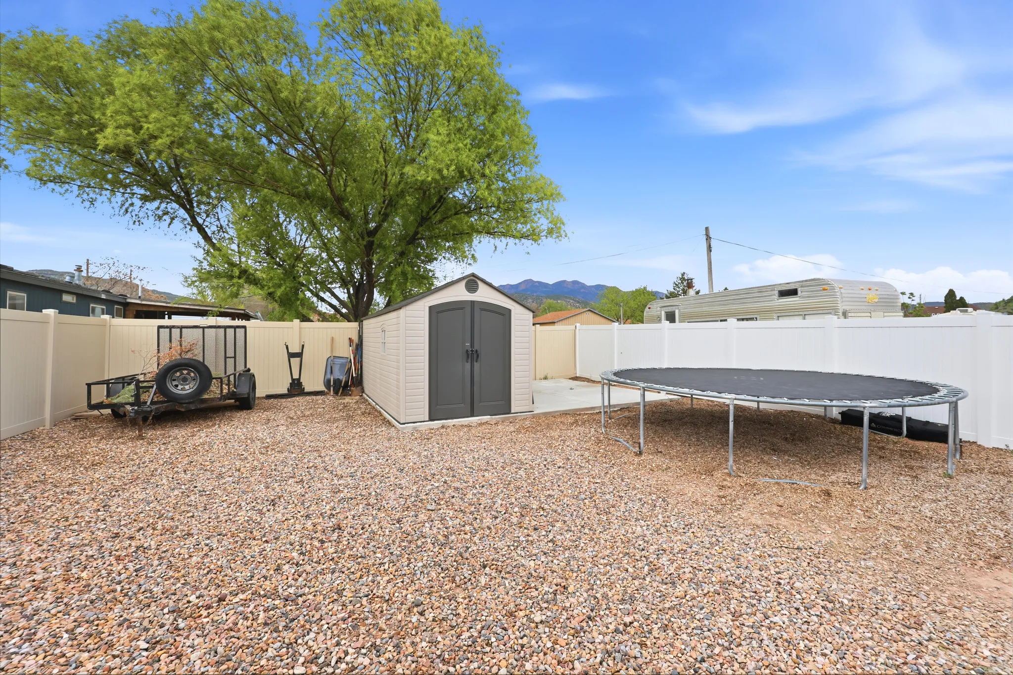 Expansive gravel yard featuring a privacy fence, a portable storage shed, a concrete pad, and mature trees