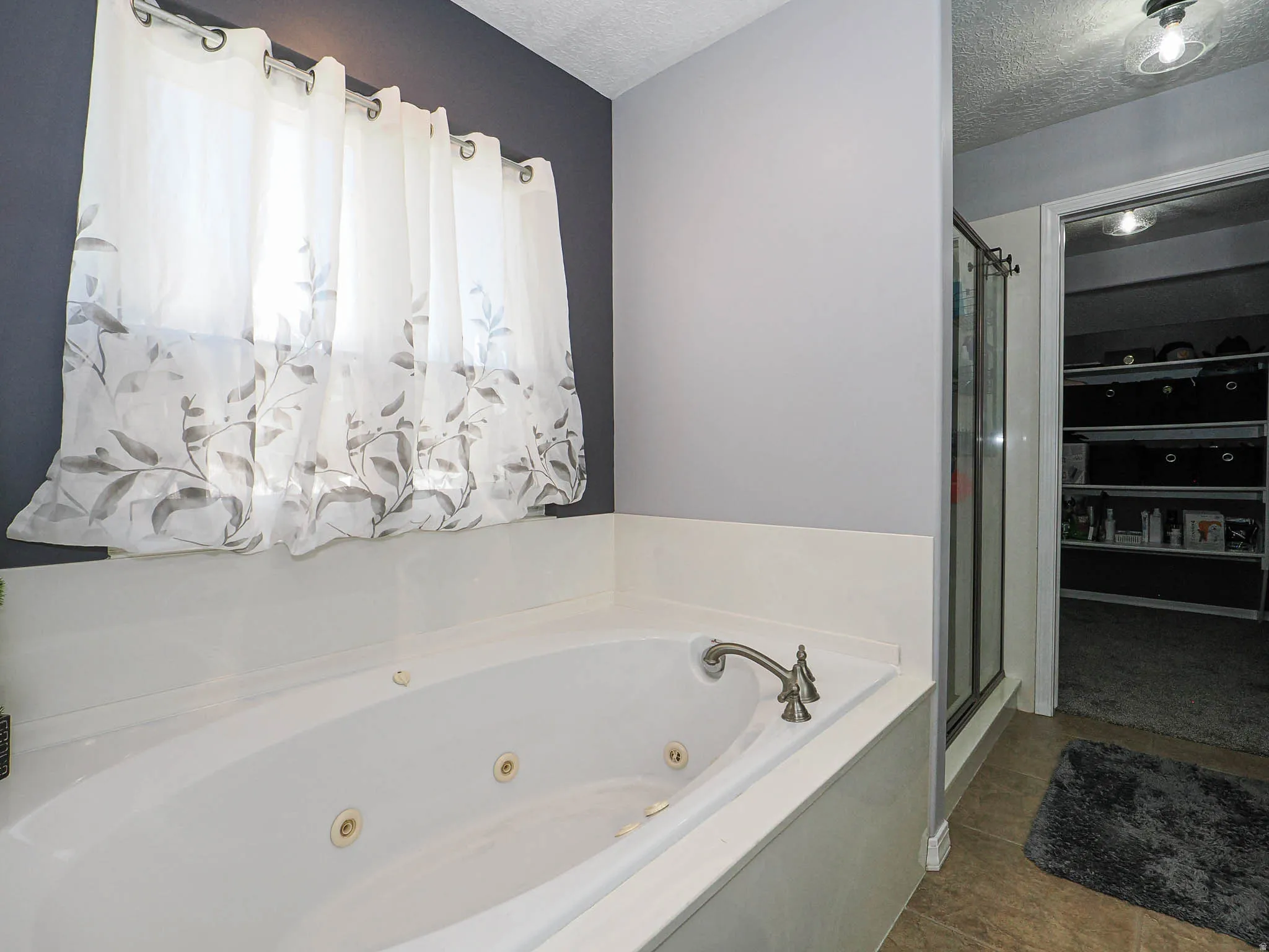 Jetted soaking tub with deck-mounted brushed nickel fixtures, under a window with sheer curtains
