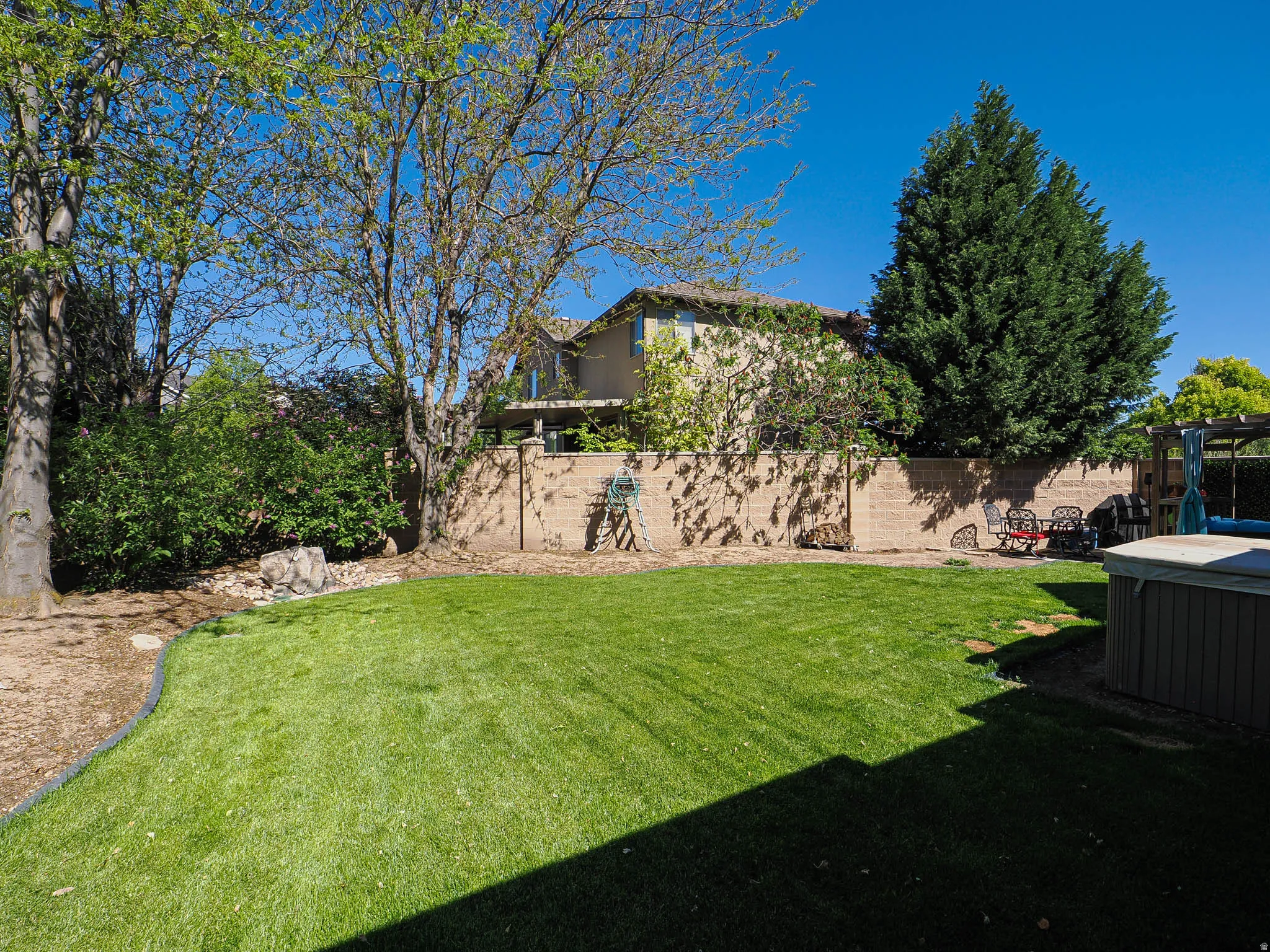 Expansive backyard featuring a manicured lawn, mature trees, a concrete block privacy wall, and a built-in spa