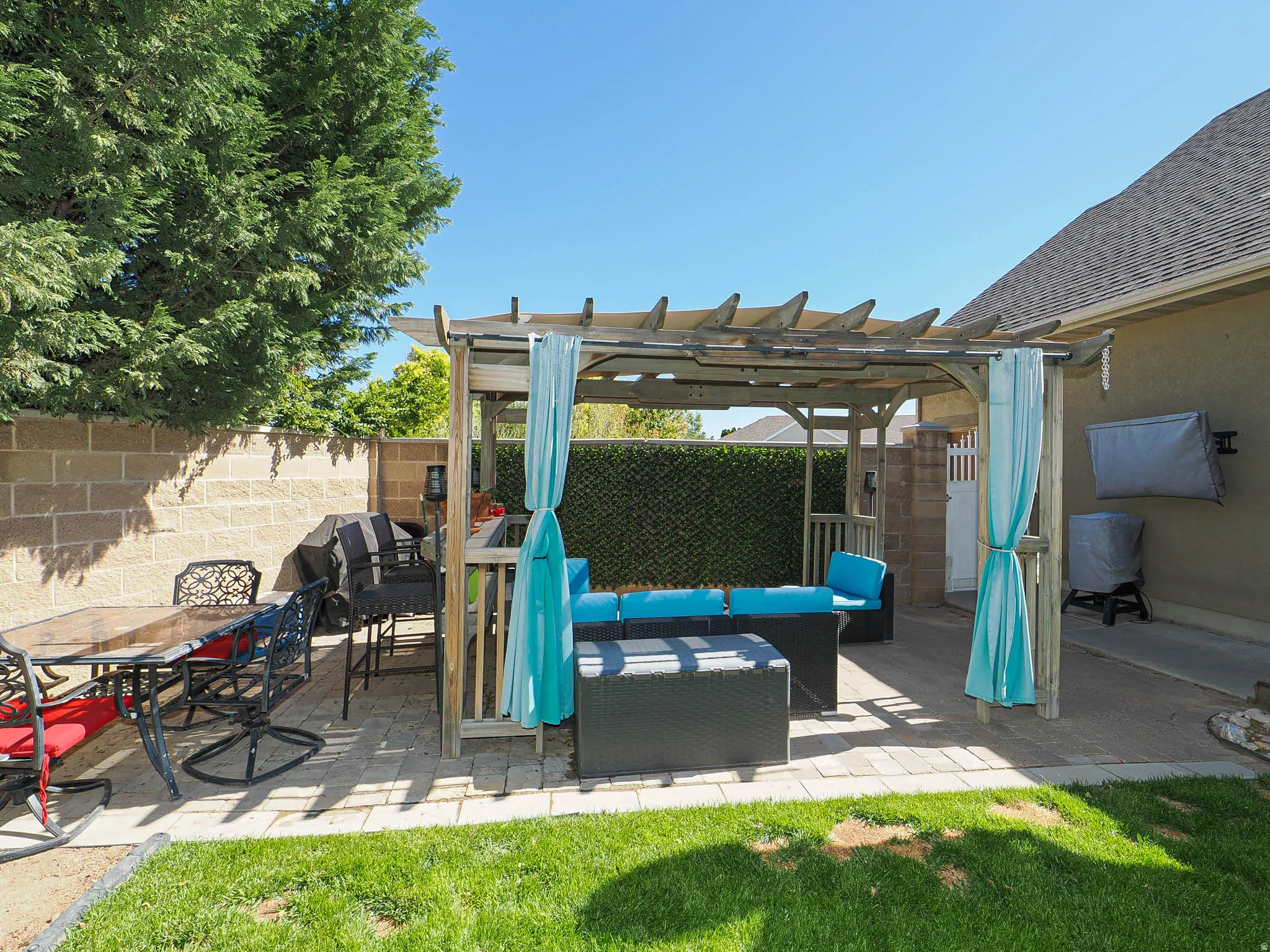 Paved patio area featuring a wooden pergola with canvas shading, a privacy hedge, and a block wall perimeter