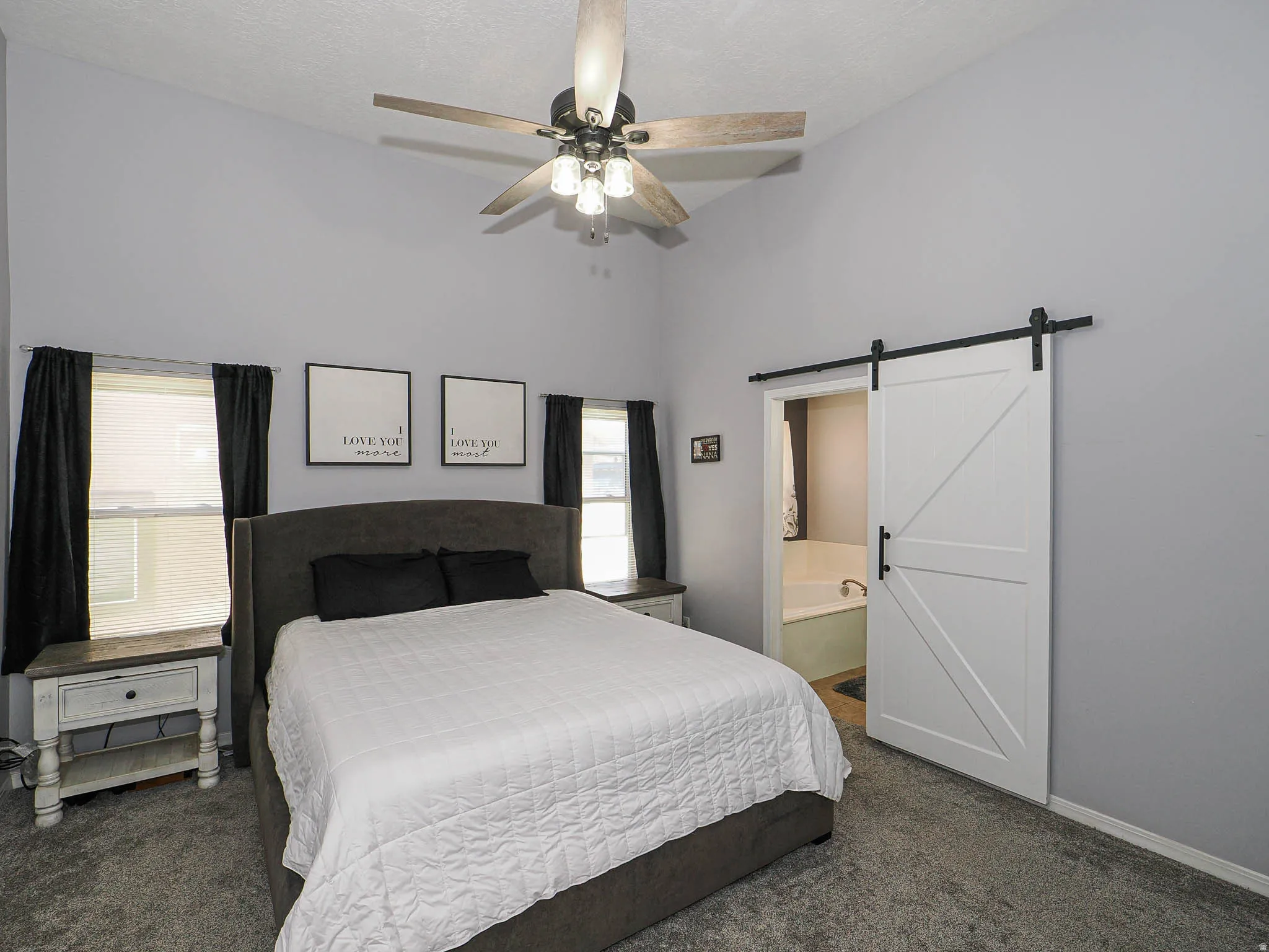 Vaulted ceiling bedroom featuring a ceiling fan with light kit, twin windows with blinds, plush gray carpeting, and a white barn door with black hardware opening to a bathroom with a built-in tub