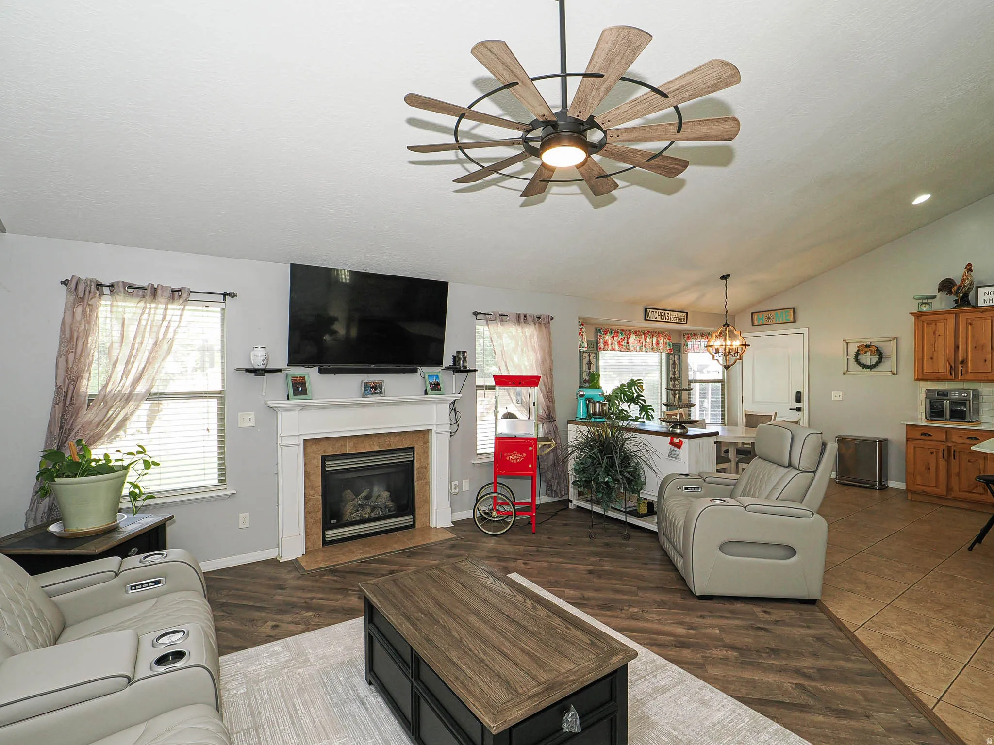 Spacious living area featuring wood-finish flooring, a white mantel fireplace with tile surround, and vaulted ceilings with a unique ceiling fan fixture