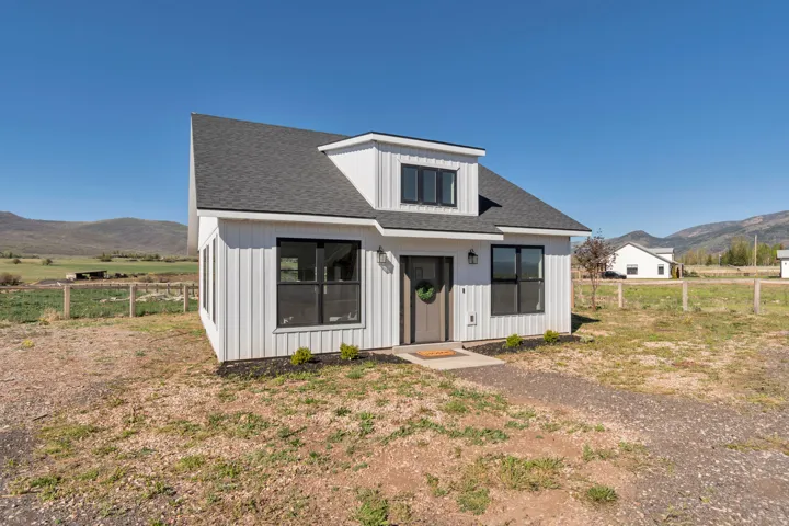 Modern farmhouse-style exterior featuring vertical white siding, black-framed windows, a dark shingled roof, a dormer window, and a clear mountain backdrop