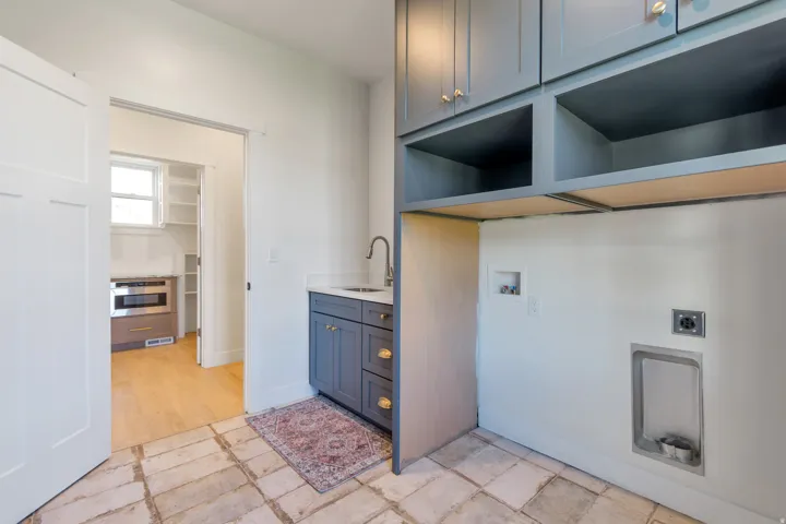 Dedicated laundry area featuring slate blue cabinetry with gold-tone hardware, a sink with a gooseneck faucet, and built-in shelving