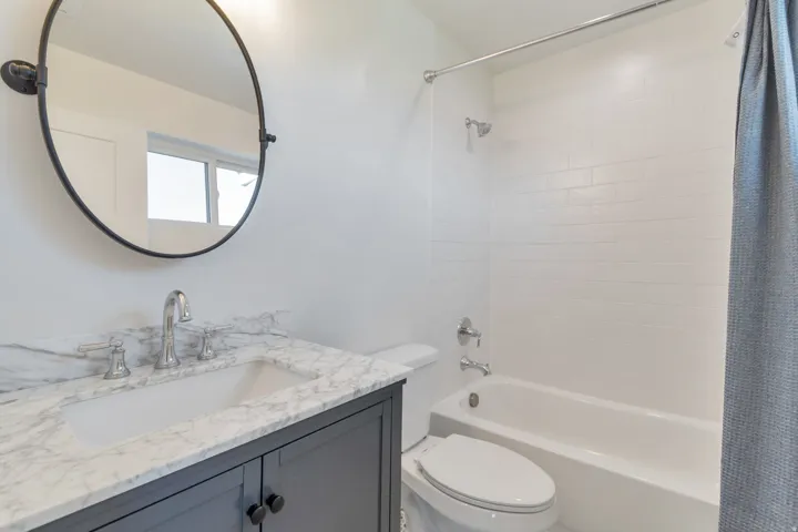 Bathroom featuring a dark grey vanity with a white countertop and undermount sink, a round black framed mirror, a white toilet, and a white tiled shower-over-tub combination with chrome fixtures