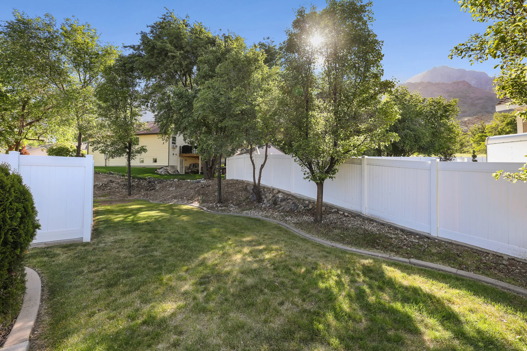 Expansive backyard featuring a manicured lawn, white privacy fencing, mature trees, and a mountain backdrop