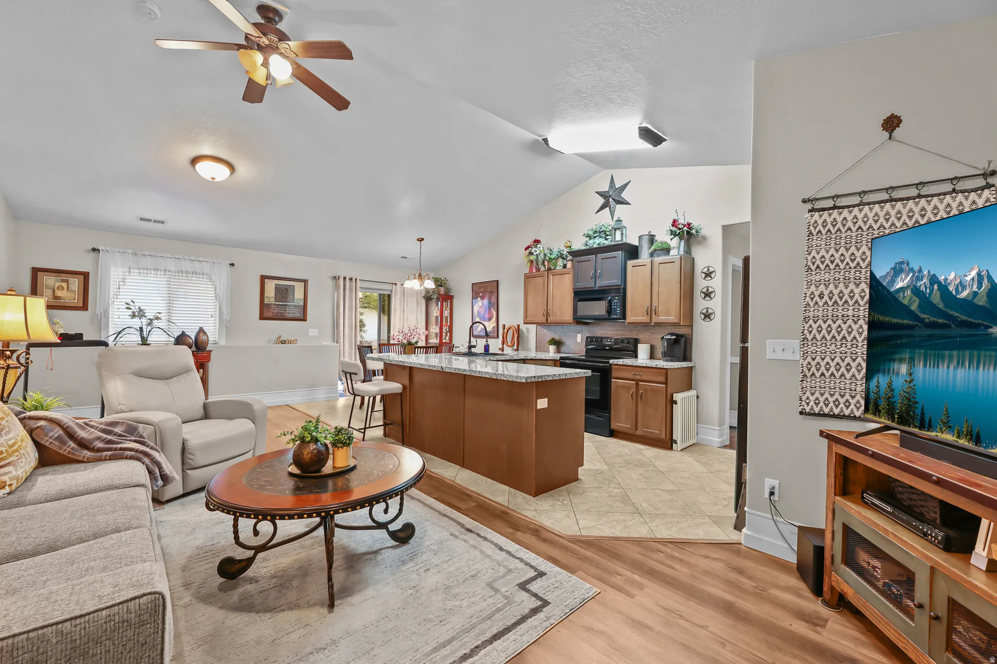 Open-concept living area featuring vaulted ceilings, a ceiling fan with integrated lighting, wood-finish flooring, and a kitchen with an island and granite countertops