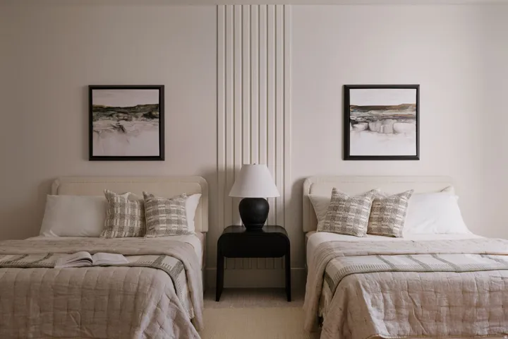 Bedroom featuring two beds with upholstered headboards, a central fluted wall panel, and a dark wood side table with a lamp