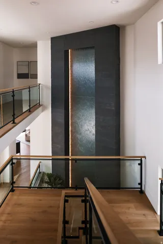 Multi-level interior featuring a stone-clad water feature with integrated lighting, glass railings with wood-finish tops, and light wood-finish flooring