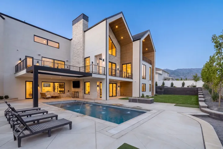 Modern stucco exterior featuring a tiered backyard with a swimming pool, stone accent chimney, multiple balconies with black railings, and an integrated pergola