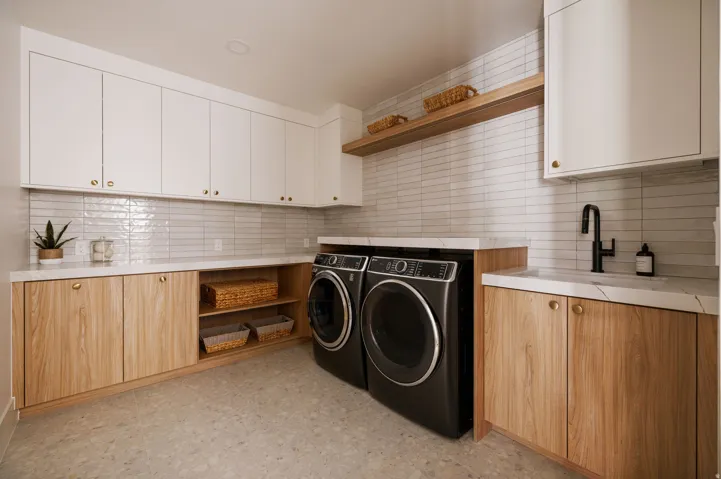 Modern laundry space featuring white upper cabinetry, wood-finish lower cabinetry, light stone countertops, and a full-height stacked tile backsplash